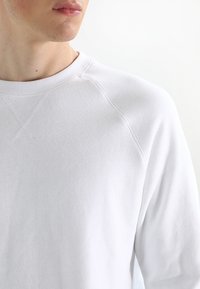 Sudadera blanca con cuello redondo, confeccionada en tela suave, con puños y dobladillo acanalados, y detalles de costuras visibles en las uniones de los hombros.