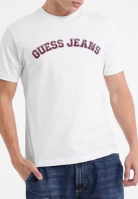 Balta medvilninė marškinėliai su ryškiu juodu ir raudonu užrašu "GUESS JEANS", arkos forma, apvaliu iškirpimu, trumpomis rankovėmis ir laisvu siluetu.
