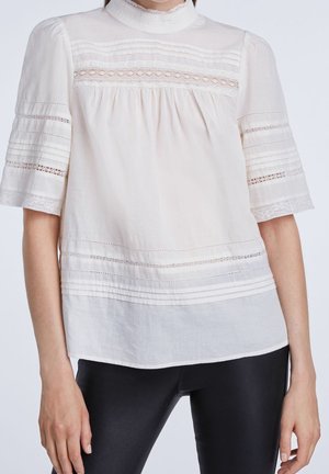 Femme portant un chemisier blanc ample avec des détails en dentelle et plissés sur les manches, le décolleté et l'ourlet, associé à un pantalon noir ajusté.