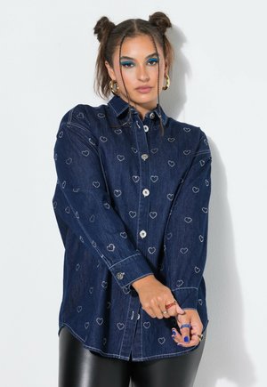 Frau mit zwei Dutfrisuren und blauem Lidschatten, die ein dunkelblaues Jeanshemd mit weißen Herzmustern und schwarze Hosen trägt, zieht ihren Ärmel zurecht.