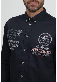 Marineblå skjorte med knapper laget av bomull. Har broderte tekster: "924 REG. TM," "FQ1924," og "Authentic PERFORMANCE." Hvite knapper.