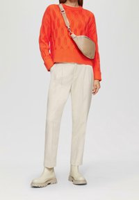 Leuchtend orangefarbener gestrickter Pullover mit strukturiertem Rautenmuster, beiger Umhängetasche, hellbeige maßgeschneiderte Hose und klobigen cremfarbenen Stiefeln.