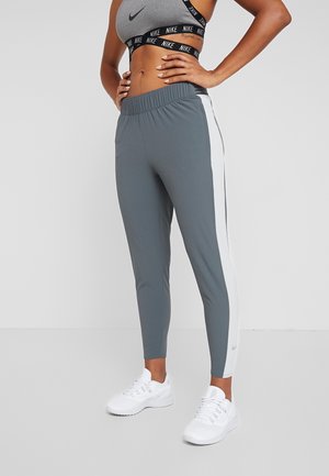 Frau trägt graue und weiße sportliche Leggings, einen grauen Nike-Sport-BH und weiße Sneakers, steht mit den Händen in die Hüften vor einem einfarbigen Hintergrund.