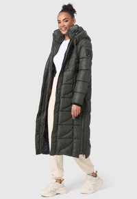 Lange donzen jas in donkergroen, met een gewatteerd ontwerp, met capuchon, rits en een rode voering. Gecombineerd met beige sweatpants en witte sneakers.