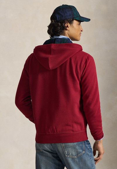 Sweatshirt com capuz vermelho feito de material macio, com bolso canguru e punhos canelados. Usado com jeans azuis e um boné xadrez.