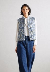 Γιλέκο με λουλουδάτο σχέδιο σε ανοιχτό μπλε χρώμα, με στρογγυλή κολαρία, κλείσιμο με κουμπιά και δύο μπροστινές τσέπες, φορεμένο πάνω από λευκή μπλούζα και μπλε τζιν παντελόνι.