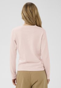 Dolga majica svetlo roza barve iz mehke tkanine, z okroglo izrezom in preprostim dizajnom, v kombinaciji z bež hlačami.
