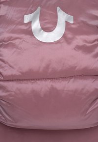 Růžová polstrovaná bunda s lesklou hladkou texturou a velkým bílým logem ve tvaru U na přední straně. Výrazné švy jsou viditelné.