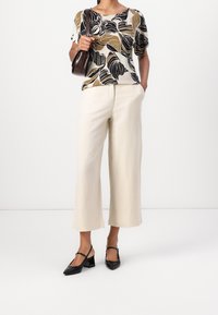 Haut à manches courtes, coupe décontractée, avec un motif de feuilles en noir, marron et crème. Associé à un pantalon large crème et des chaussures à talons ouverts noires.