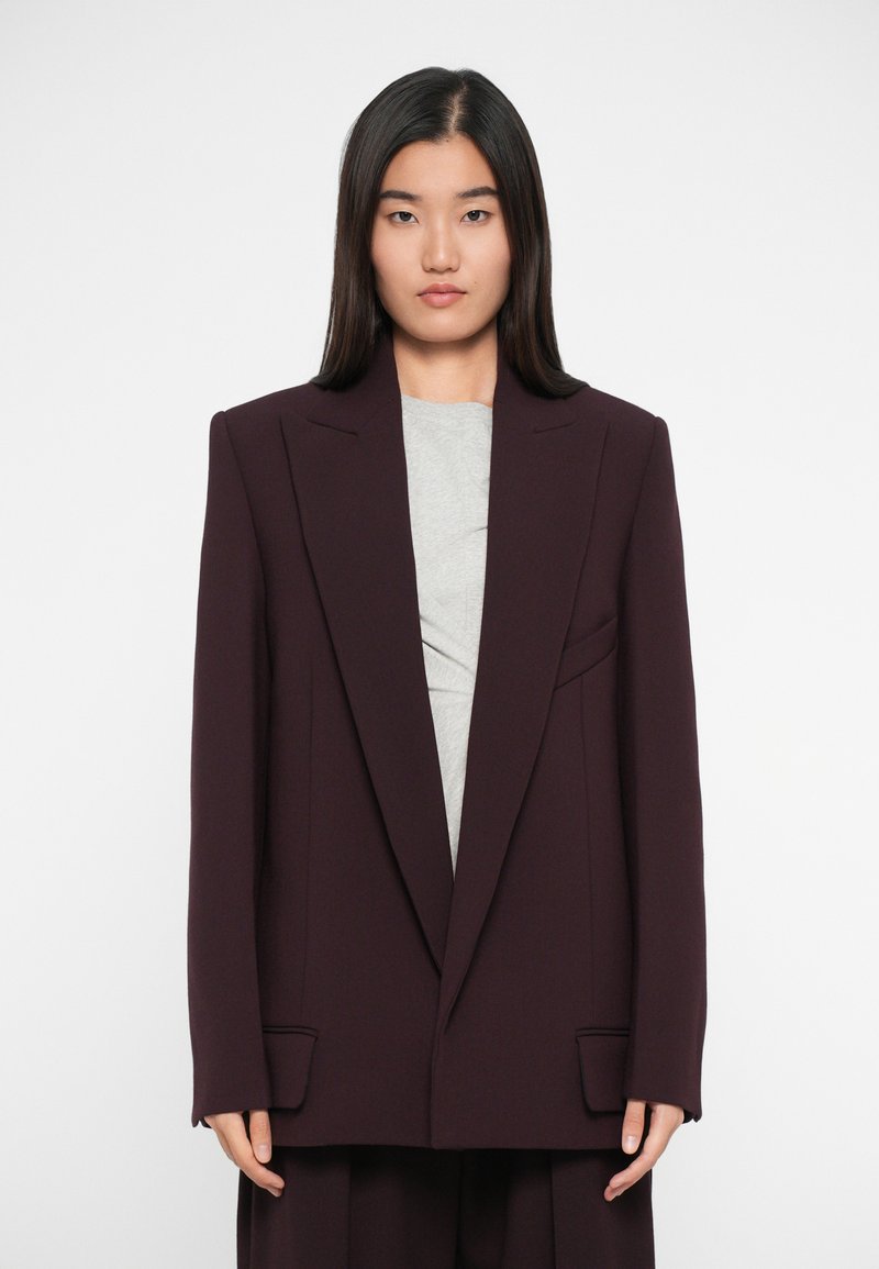 Burgundský oversize blazer s tvarovanými rameny, klopovými límci a dvěma předními kapsami, navrstvený na šedém topu. Hladká textura tkaniny.