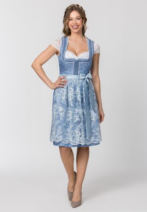 Frau lächelt, trägt ein blaues traditionelles Kleid mit weißer Spitzen-Schürze und kurzen Spitzenärmeln, steht mit einer Hand in der Hüfte und beigen Absätzen.