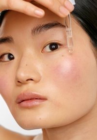 En klar pipettflaska som släpper ut ett serum, hålls över en models kind med en subtil rosa nyans. Huden är len med en naturlig lyster.