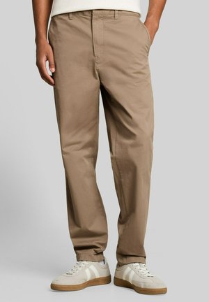 Homme portant un pantalon beige décontracté et des baskets blanc cassé, debout les mains dans les poches devant un fond uni.