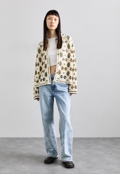 Heklet cardigan i krem- og grønnmønster, kombinert med lyseblå jeans og svarte sko. Har løs passform og åpen frontdesign.