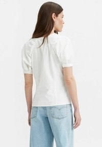 Blusa bianca in cotone con maniche corte a sbuffo e dettaglio arricciato sul retro, abbinata a jeans in denim azzurro chiaro. Design semplice e pulito.