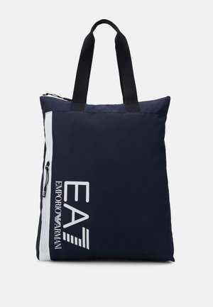 Borsa tote blu navy con manici neri, fascia verticale bianca, tasca con cerniera e grande logo bianco "EA7 Emporio Armani" sulla parte frontale.