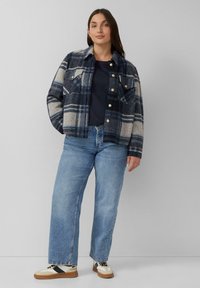 Karierte Flanelljacke in Navy- und Beigetönen, mit zwei vorderen Taschen, Druckknöpfen und einer lässigen Passform, kombiniert mit hellblauen Jeans und Sneakers.
