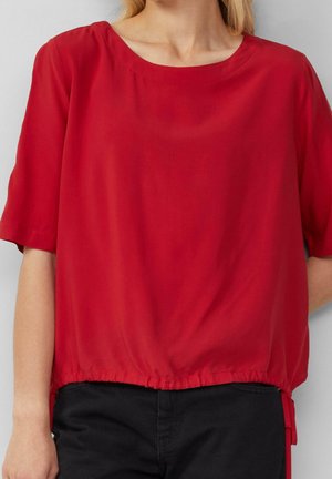 Czerwona bluzka z krótkim rękawem wykonana z lekkiego materiału. Posiada zaokrąglony dekolt i marszczony detal na dole, zestawiona z czarnymi spodniami.