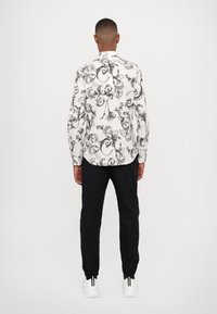 Camisa blanca de manga larga con un patrón ornamental negro, combinada con pantalones negros y calzado blanco con acentos negros.
