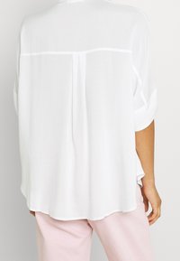 Blusa bianca ampia con orlo arrotondato, maniche corte arrotolate e una sottile plissettatura sulla schiena. Il tessuto appare leggero e liscio.