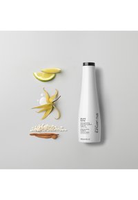 Bouteille de shampoing blanche avec un capuchon noir, étiquetée "izumi tonic", entourée de tranches de citron, de pétales de fleurs, de grains et d'un bâton en bois sur un fond gris.