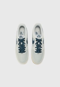 Nike Air Force 1 -lenkkarit vaaleanharmaasta nahasta, tummansinoin yksityiskohdin ja pyörillä varustetuin nauhoin. Rei'itetty varvasosa ja kumipohja.