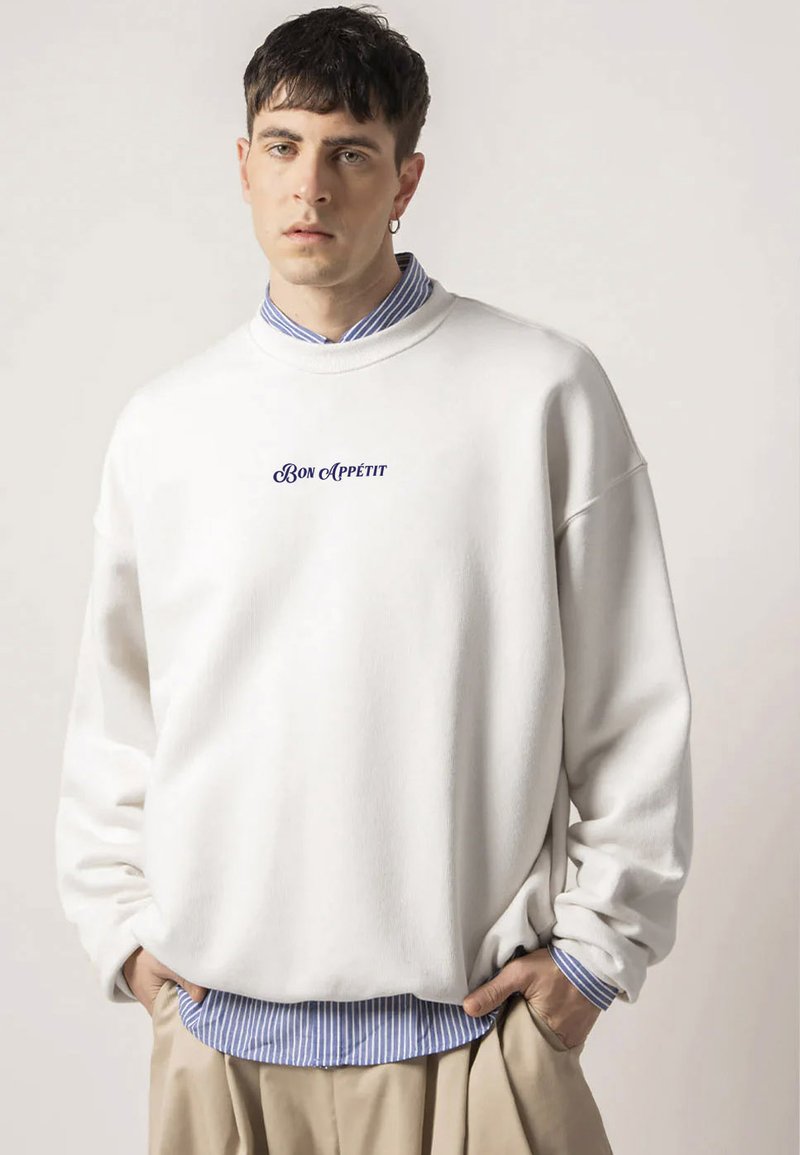 White sweatshirt with a round neck, featuring "Bon Appétit" in navy cursive. Worn over a blue and white striped shirt, textured fabric.