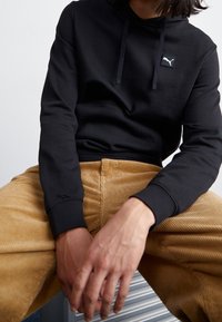 Persona che indossa un hoodie nera Puma e pantaloni in corduroy beige, seduta con le mani intrecciate tra le gambe su uno sfondo semplice.