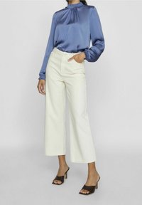 Hellblaue Satinbluse mit hohem Kragen und elastischen Bündchen, kombiniert mit cremefarbenen weit geschnittenen Hosen; Modell trägt braune Absatzmules.