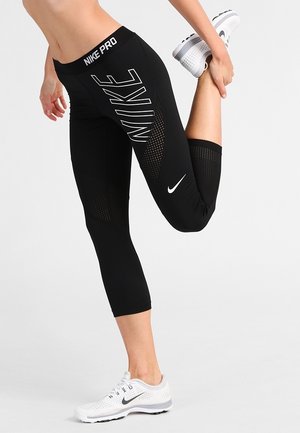 Osoba wyciągająca nogę do tyłu, ubrana w czarne legginsy Nike Pro z białymi logo oraz białe buty do biegania marki Nike na jednolitym tle.
