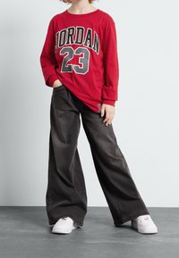 Rød langærmet skjorte med "JORDAN 23" grafik; kombineret med løstsiddende, mørke denim bukser og hvide sneakers. Afslappet, komfortabelt tøj.