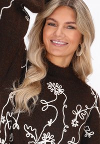 Pulover tricotat maro, cu broderie florală albă, având guler înalt și mâneci lungi. Textură netedă și croială lejeră.