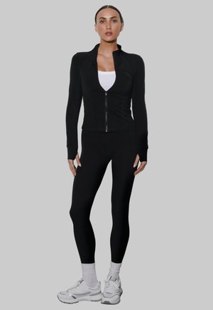 Kobieta stojąca w czarnej kurtce na zamek błyskawiczny, czarnych legginsach, białej podkoszulce, białych skarpetach i białych trampkach na szarym tle.