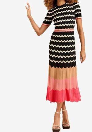 Femme portant une robe midi à blocs de couleurs avec motif zigzag en noir, blanc, beige, rose et rouge, ainsi que des sandales à talons hauts beiges.