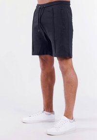 Zwarte shorts met een trekkoord in de taille, zijzakken en een gestructureerd logo-detail op het been. Gepaard met witte sneakers.