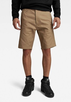 Pantalones cortos caqui con un frente plano, bolsillos laterales y un corte entallado. Llevados con zapatillas negras de caña alta que cuentan con detalles de tiras y logotipos.
