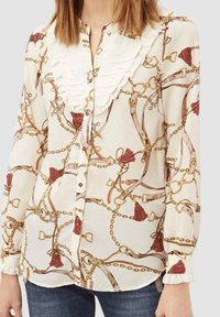 Blusa blanca de botones con cuello con volantes, patrón de cadenas y flecos en rojo y dorado, con una textura de tela suave y puños fruncidos.