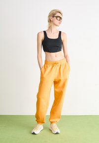 Top corto negro y pantalones deportivos naranjas brillantes con puños elásticos. Zapatillas blancas con un diseño texturizado y acentos amarillos. Suelo verde por debajo.