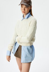 Crème kabelgebreide trui met ribbelzoom, gelaagd over een lichtblauw overhemd met knoopjes en een gemarmerde bruine short. Accentuatie met een bruine leren pet.