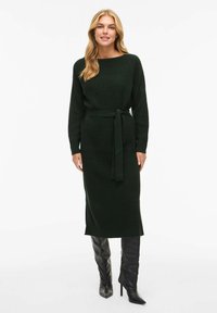 Ciemnozielona dzianinowa sukienka z długimi rękawami, okrągłym dekoltem i paskiem w talii. Charakteryzuje się dopasowaną sylwetką i długością do połowy łydki. Stylizowana z kozakami za kolano.