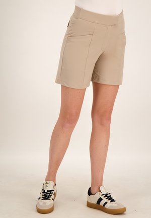 Persoon draagt beige knielange shorts en witte sneakers met zwarte strepen, staand tegen een effen lichte achtergrond.