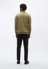 Olívazöld pulóver gallérral, bordázott mandzsettákkal és szegéllyel, fekete nadrággal és cipővel párosítva, hátulnézetben.