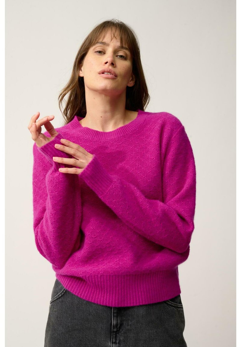 Pull en laine rose vif avec une finition texturée, doté d'un col rond, de poignets côtelés et d'une ceinture côtelée. Associé à un jean foncé.