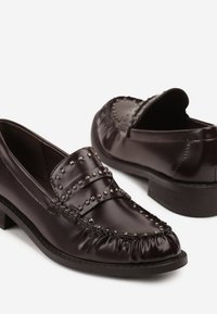 Mocassins en cuir marron foncé avec des accents cloutés, dotés d'un bout arrondi, d'un dos élastiqué et d'un petit talon carré. Texture lisse avec un détail froncé.