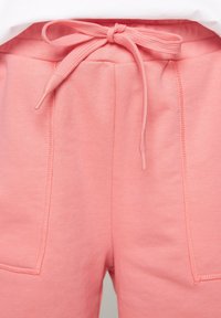 Pantaloni della tuta rosa chiaro con cintura regolabile, dotati di due tasche frontali e cuciture in vista. Texture morbida con un design casual.