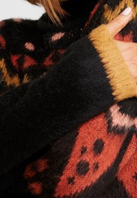 Pull noir en maille avec des motifs orange, rouge et jaune. Dispose d'un poignet côtelé moutarde et d'un tissu doux et texturé.
