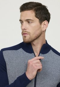 Pull zippé bleu marine et gris avec un tricot texturé, présentant une coupe ajustée et un col montant, mettant en valeur des blocs de couleurs contrastantes.