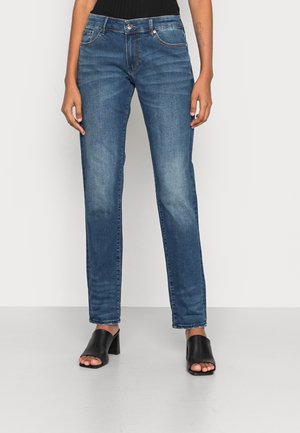 Person iført blå straight-leg jeans med middel talje og falmede detaljer samt sorte åbne blokhæls-sandaler, stående mod en ensfarvet baggrund.