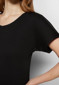 Gros plan d'une personne portant un haut noir côtelé à manches courtes ajusté, main posée sur la hanche, peau claire et cheveux longs bruns.