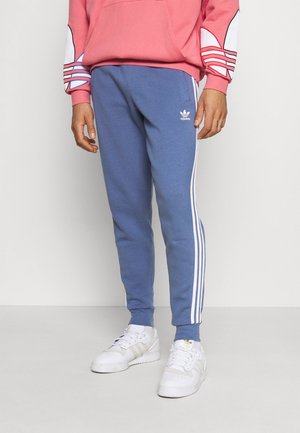 Mężczyzna noszący niebieskie spodnie dresowe Adidas z białymi paskami i białe trampki, w połączeniu z różową bluzą z logo na rękawach.